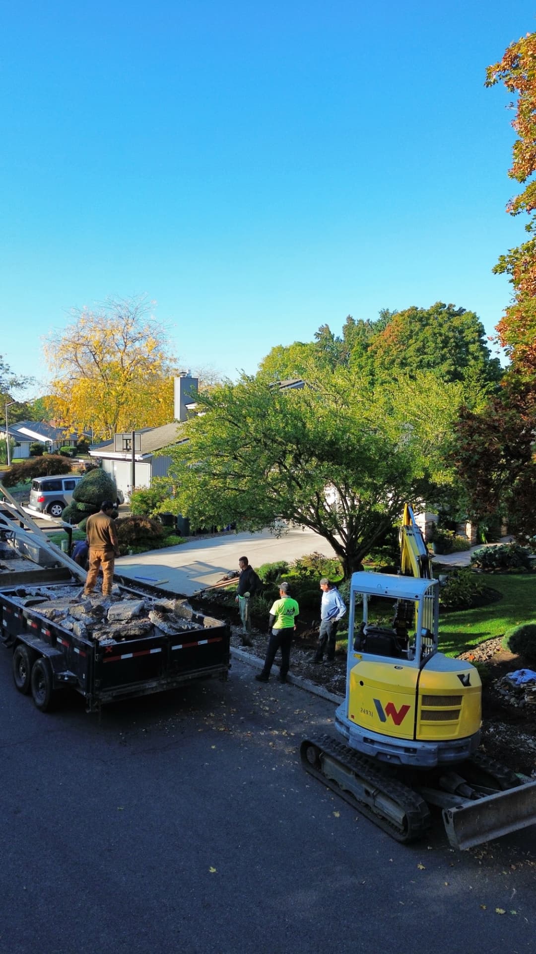 Aerial view of a driveway replacement under way, with a mini-excavator and a trailer loaded with broken concrete beside a suburban home